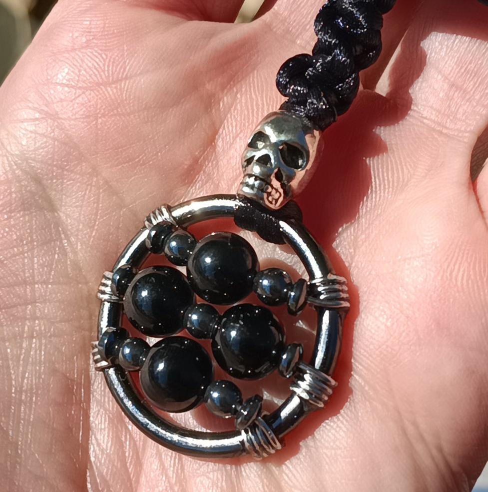 Fidget necklace consisting of a gunmetal grey round metal ring with three wires strung across it. ad wrapped around it. the fidget mechanism consists of 4 large black tourmaline beads in a plus shape, with one centered on the top wire, two on the middle wire, and one centered on the bottom wire, interspersed with small hematite beads. The fidget is a pendant on a black nylon cord, with a silver skull shaped bead strung onto the cord atop the fidget, which is topped with a section of black braided nylon.