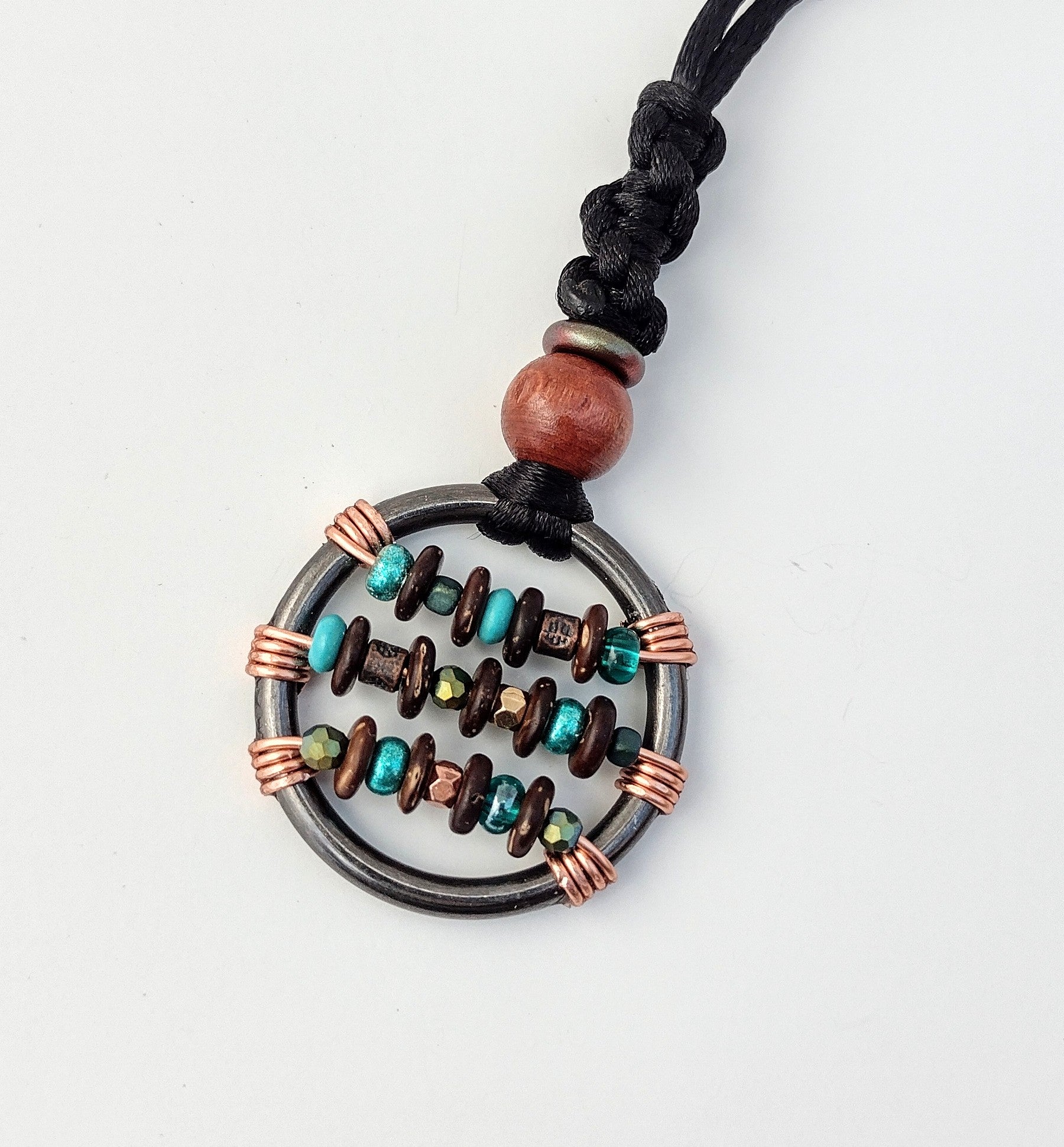 a 1.25 inch round dark gray metal ring, with three copper wires wrapped across it horizontally, on the wires are small beads, teal glass, turquoise, copper, and iridescent green, alternating with small flat wooden beads. The pendant is hung on a sleek black nylon cord, with a round wooden bead, a copper colore bead, and a section of black braiding securing the fidget.